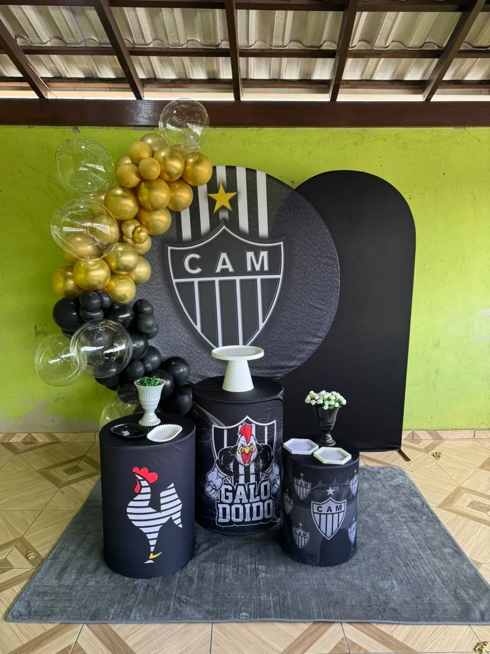 Decoração Completa com tema do Atlético Mineiro: painel redondo temático e painel arqueado preto, arco desconstruído de balões dourados, pretos e bubbles transparentes, trio de cilindros com capas do tema, boleira, bandejas, vasos de flores e tapete