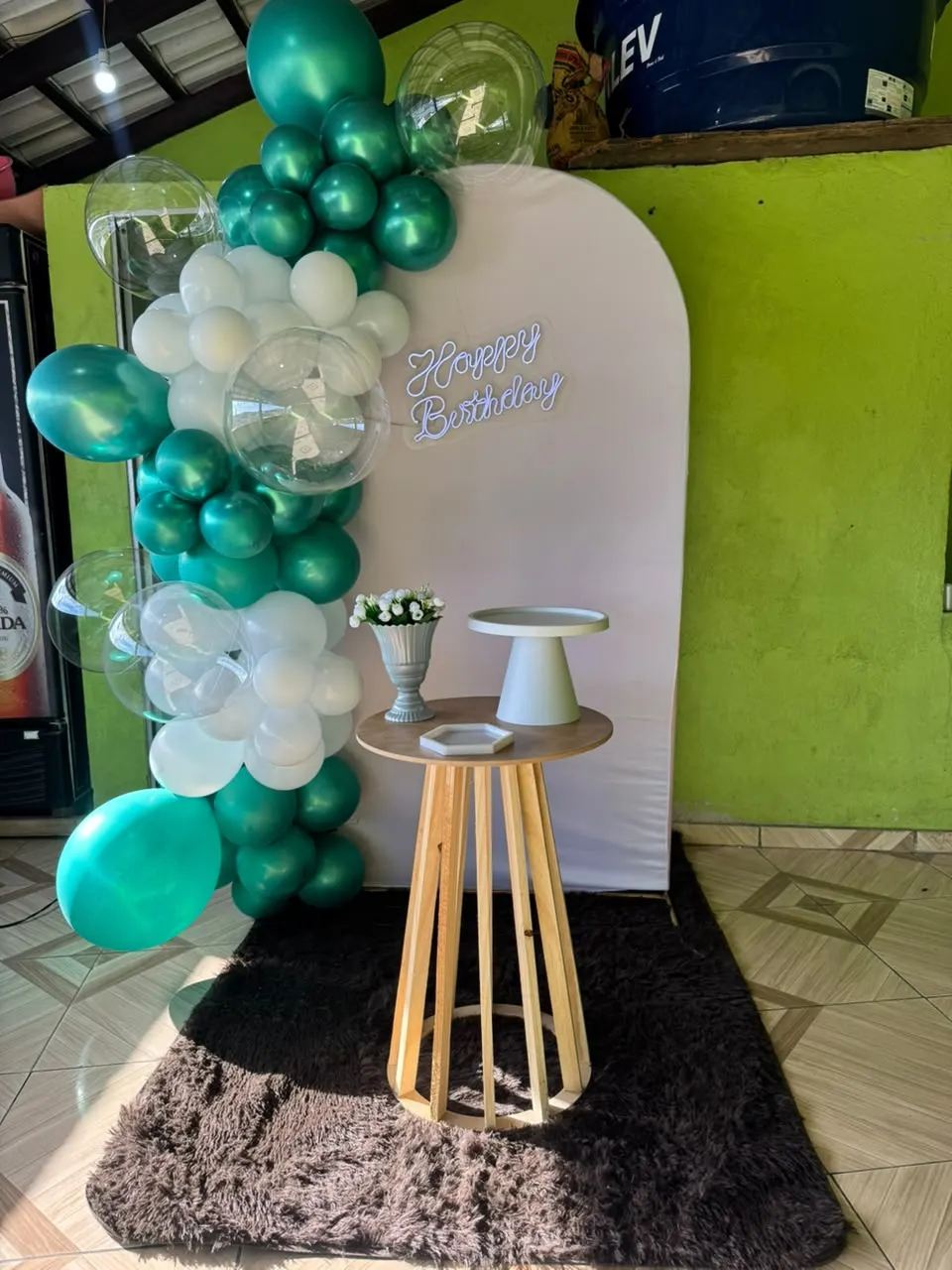 Decoração Essencial: painel em arco branco com letreiro neon Happy Birthday, arco desconstruído de balões verdes, brancos e bubbles transparentes, mesa cilíndrica ripada de madeira clara com boleira, bandeja e vaso de flores, sobre tapete felpudo marrom