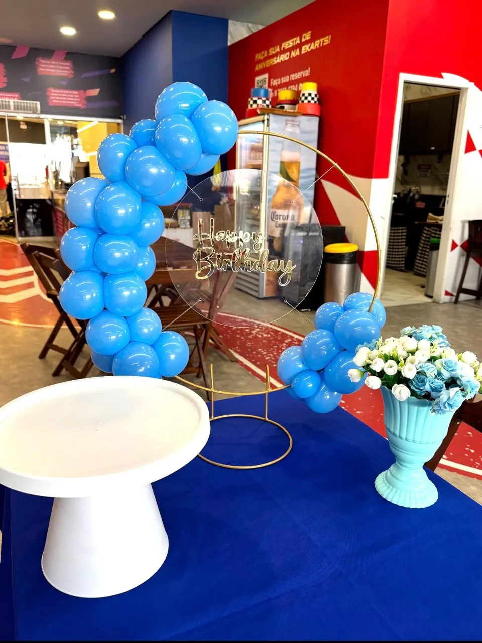 Kit Festa na Mesa: mini painel redondo Happy Birthday com arco de balões azuis, boleira branca e vaso de flores sobre a mesa
